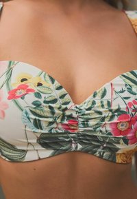 Bikini gornji dio s cvjetnim uzorkom u bijeloj boji s ružičastim, žutim i zelenim motivima; ima nabranu konstrukciju i izrez u obliku srca.