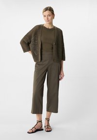 Brauner, offener Strickcardigan über einem figurbetonten grünen Oberteil, kombiniert mit olivgrünen, weiten Hosen und schwarzen Sandalen mit Riemchen. Einfacher, geschichteter Look.