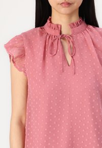 Blusa rosa chiaro con scollatura arricciata e dettaglio con fiocco. Presenta pois testurizzati su tutta la superficie e maniche corte svasate per un volume extra.