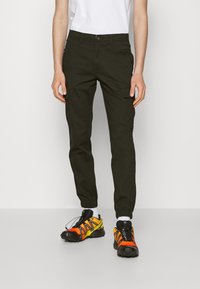 Oliwkowe spodnie typu cargo ze zwężanymi mankietami, wyposażone w liczne kieszenie i zapięcie na guziki, zestawione z jaskrawo pomarańczowo-czarnymi butami sportowymi.