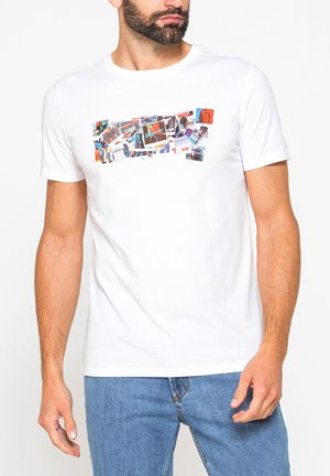 Uomo con la barba che indossa una t-shirt bianca a maniche corte con stampa colorata a collage fotografico rettangolare e jeans blu, in piedi su sfondo bianco.