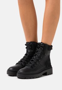 Bottes de combat en cuir noir à lacets avec des semelles épaisses à crans, portées sur des jambes nues sur fond blanc.