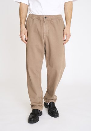 Mand iført løstsiddende beige bukser med elastisk talje og knap, kombineret med sorte læder slip-on sko og hvide sokker.