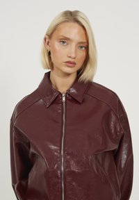 Giacca di pelle borgogna con una texture lucida, colletto ampio, chiusura lampo frontale e vestibilità oversize, con spalle scese e tasche laterali.