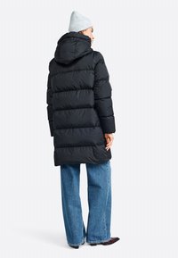 Zwarte pufferjas met een capuchon, met een gewatteerde textuur, lange lengte en zijzakken. Gecombineerd met blauwe wijde jeans en een lichtgrijze muts.