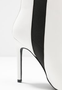 Zapato de tacón alto blanco con un panel lateral de tela negra texturizada. Presenta una punta afilada y un acabado elegante y brillante.