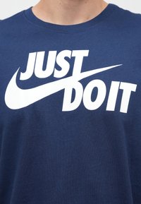 Μπλουζάκι από βαμβάκι σε ναυτικό μπλε χρώμα με μεγάλο λευκό κείμενο "JUST DO IT" και γραφικό swoosh, στρογγυλή λαιμόκοψη και κοντά μανίκια.