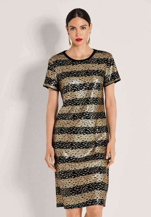 Frau mit dunklem Haar, die ein kurzärmeliges Kleid mit goldenen und schwarzen Pailletten mit horizontalen Streifen trägt, steht vor einem einfarbigen hellen Hintergrund.