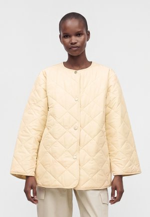 Personne portant une veste matelassée beige clair boutonnée avec de larges manches et un pantalon cargo couleur crème sur un fond uni.