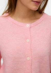 Ροζ μάλλινη ζακέτα με στρογγυλή λαιμόκοψη, κλείσιμο με πέντε κουμπιά και μαλακό, υφασμάτινο υλικό με υφή. Τα κουμπιά είναι λευκά με διακριτικό γκλίτερ.