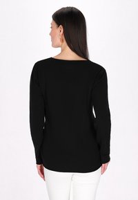 Top negru cu mânecă lungă, guler rotund, textură netedă și tiv curbat. Modelul este prezentat din spate, purtând pantaloni albi.
