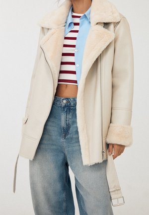 Person trägt eine beige Schaffelljacke über einem blauen Hemd mit Knöpfen und einem rot-weiß gestreiften, bauchfreien Top mit hoch taillierten blauen Jeans.