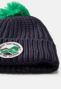 Berretto lavorato a maglia di colore blu scuro con un design a coste testurizzate, caratterizzato da un pompon verde e un patch ricamato con grafiche di montagne.