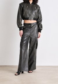 Czarna skórzana skrócona kurtka bomberka z dwoma suwakami z przodu; zestawiona z szerokimi skórzanymi spodniami typu cargo. Teksturowana tkanina i detal z paskiem.