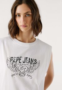 Donna con capelli castani alla spalla che indossa un top bianco senza maniche con una grafica a cuore alato nera "Pepe Jeans London".