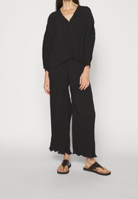 Svart texturerad outfit bestående av en lös V-ringad topp och vida byxor med fransade fållar. Bärs med platta svarta sandaler.