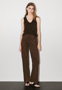 Pantaloni larghi in suede marrone abbinati a un top smanicato scuro. La modella indossa ciabatte nere. Lo sfondo è bianco uniforme.