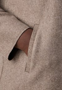 Détail d'un manteau en feutre marron avec une poche poitrine, présentant une texture textile lisse et un accent en cuir marron contrastant visible sur le bras.