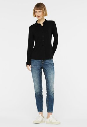 Frau steht und trägt ein schwarzes Hemd mit Knöpfen, blaue Jeans und weiße Turnschuhe vor einem schlichten weißen Hintergrund.