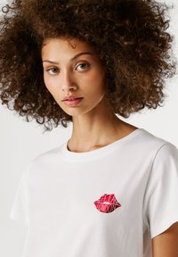 Νεαρή γυναίκα με σγουρά μαλλιά που φοράει ένα λευκό t-shirt με μια μικρή θηλυκή κουκίδα που γράφει "Pepe Jeans London".