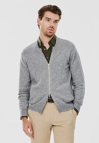 Szary kardigan na zamek wykonany z miękkiej dzianiny, z okrągłym dekoltem i ściągaczami na rękawach, nałożony na ciemnozieloną koszulę z kołnierzykiem.