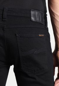 Černé džíny z denimu mají zadní kapsu s vlnitým švem, koženou záplatu na pásku a malou značkovou cedulku u kapsy.