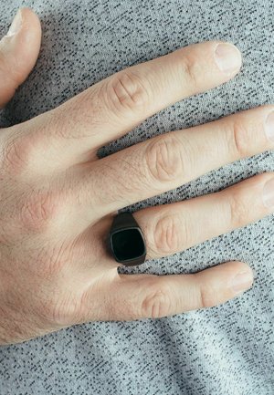 Black square signet ring on a hand, featuring a smooth, matte finish. Fingertips slightly visible against a textured gray fabric background.