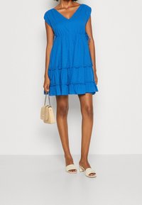 Vestido azul con volantes en capas, escote en V y mangas cortas tipo capa. Bolso de tela con correa de cadena y sandalias de tacón grueso en beige.