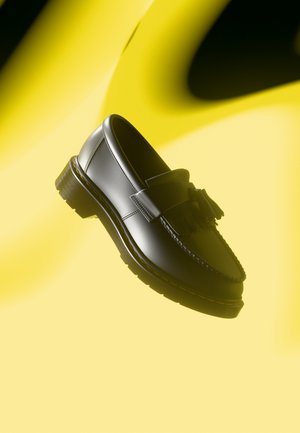 Mocasines de cuero negro con un diseño cosido, detalle de borlas y una suela texturizada, en un fondo con degradado amarillo.