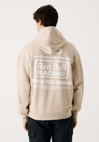 Sudadera con capucha de color beige y corte relajado, con gráficos impresos en blanco en la parte posterior, que incluyen "Red Bull Racing" y "Te Da Alas".
