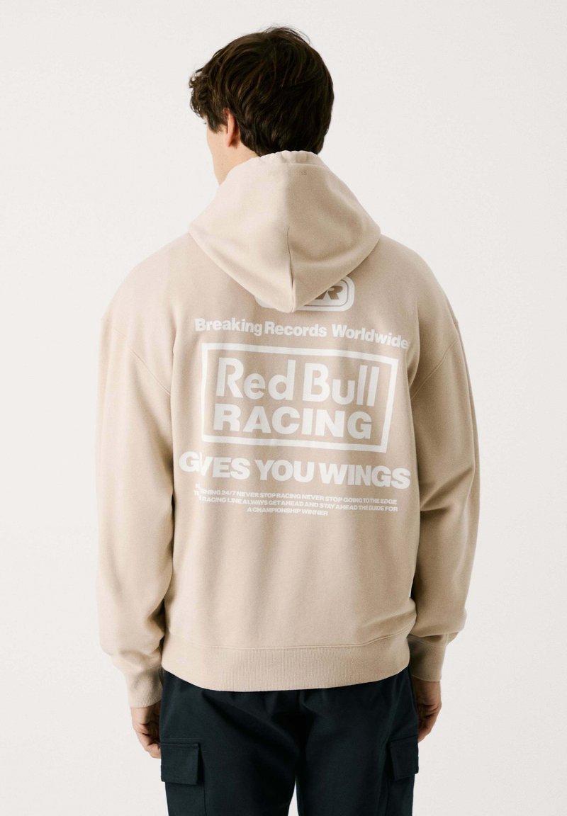 Sudadera con capucha de color beige y corte relajado, con gráficos impresos en blanco en la parte posterior, que incluyen "Red Bull Racing" y "Te Da Alas".