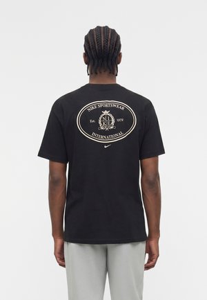 Mand med flettet hår iført sort Nike T-shirt med beige rundt "Nike Sportswear International" logo på bagsiden, lysegrå bukser, vender væk.