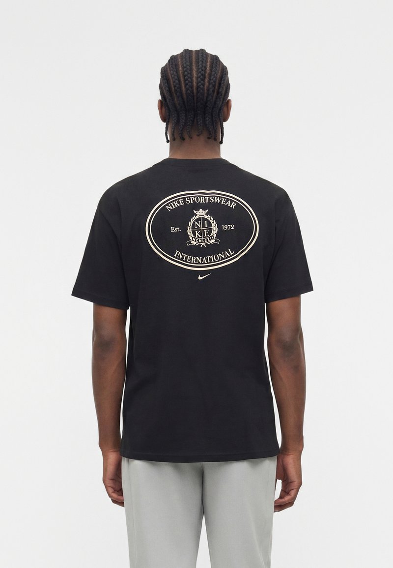 Uomo con capelli intrecciati che indossa una t-shirt nera Nike con logo circolare beige "Nike Sportswear International" sulla schiena, pantaloni grigio chiaro, di spalle.