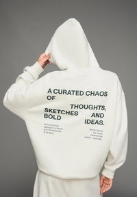 Persoon in een witte hoodie met tekst op de achterkant: "Een gecureerde chaos van gedurfde schetsen, gedachten en ideeën" en kleinere motiverende zinnen.