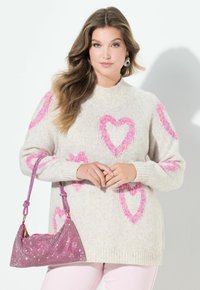 Frau mit langen, welligen Haaren, die einen beigen Pullover mit pinken Herzmustern trägt und eine funkelnde pinke Handtasche hält, steht vor einem weißen Hintergrund.