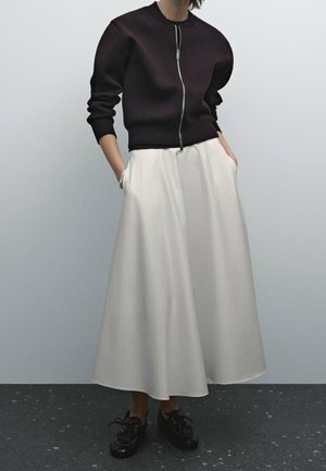 Black zip-up sweatshirt with ribbed cuffs, paired with a flowy white ankle-length skirt featuring side pockets, and black lace-up shoes.