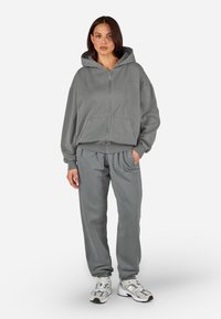 Szürke zipzáras kapucnis pulóver nagy elülső zsebekkel, szürke melegítőnadrággal párosítva. Mindkettő laza szabású és bordás mandzsettákkal rendelkezik a kényelem érdekében.