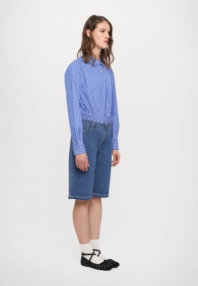 Blå och vit randig långärmad skjorta, knuten vid midjan, som är parat med knälånga denimshorts och svarta skor med prickar.
