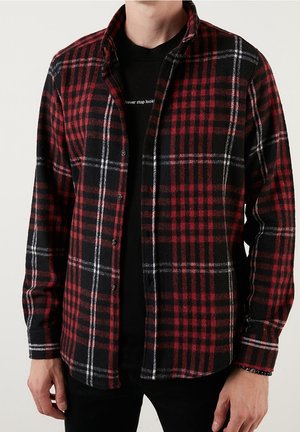 Homme portant une chemise à carreaux rouge, noire et blanche déboutonnée sur un t-shirt noir avec un texte blanc partiellement visible.