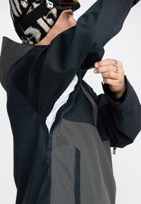 Veste de ski technique en noir et gris. Comprend un maillage blanc respirant sous les bras, des poches zippées et un extérieur imperméable.