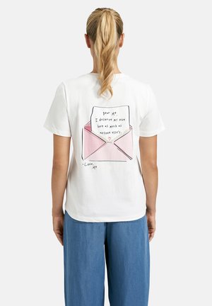 Woman with blonde ponytail wearing white t-shirt showing pink envelope illustration and letter reading self-love message on back.