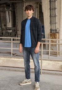 Cárdigan de punto azul marino, camiseta azul claro y jeans azules desgastados. El calzado presenta una combinación de colores gris y blanco con detalles texturizados.