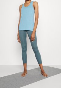 Ljusblå tanktop med rundad halsringning och sidoklipp, parat med turkosa leggings. Tyget verkar slätt och elastiskt.