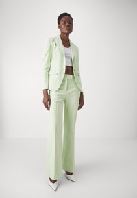Blazer su misura verde chiaro e pantaloni coordinati, caratterizzati da un design aderente e una texture liscia. Tacchi a punta bianchi completano il look.