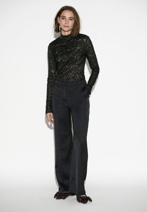 Zwarte kanten top met lange mouwen en hoge hals, gecombineerd met strakke zwarte satijnen broek. Opvallende textuur en silhouet, elegante pasvorm en subtiele glans.