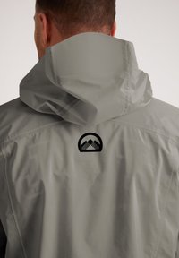 Giubbotto impermeabile grigio chiaro con grande cappuccio, texture liscia e logo nero di una montagna sul retro. Design senza cuciture con dettagli minimi.