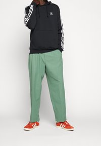 Schwarzer Kapuzenpullover mit weißen Streifen an den Ärmeln, grüne plissierte Hose und rote Sneakers mit weißen Streifen. Der Kapuzenpullover hat eine Fronttasche und ein Logo.