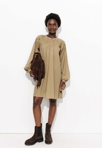 Femme portant une robe beige à manches longues avec une encolure smockée, tenant un sac à franges marron foncé et portant des bottes cheville marron foncé, se tenant contre un mur blanc.