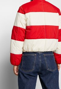 Giacca imbottita a righe rosse e crema con colletto rotondo, orlo arricciato e cuciture a vista, indossata sopra jeans in denim blu scuro.