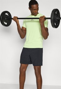 Λαχανί εφαρμοστό πουκάμισο και μαύρο σορτς. Το πουκάμισο φέρει το λογότυπο Nike Pro, και το άτομο σηκώνει μπάρα με βάρη.